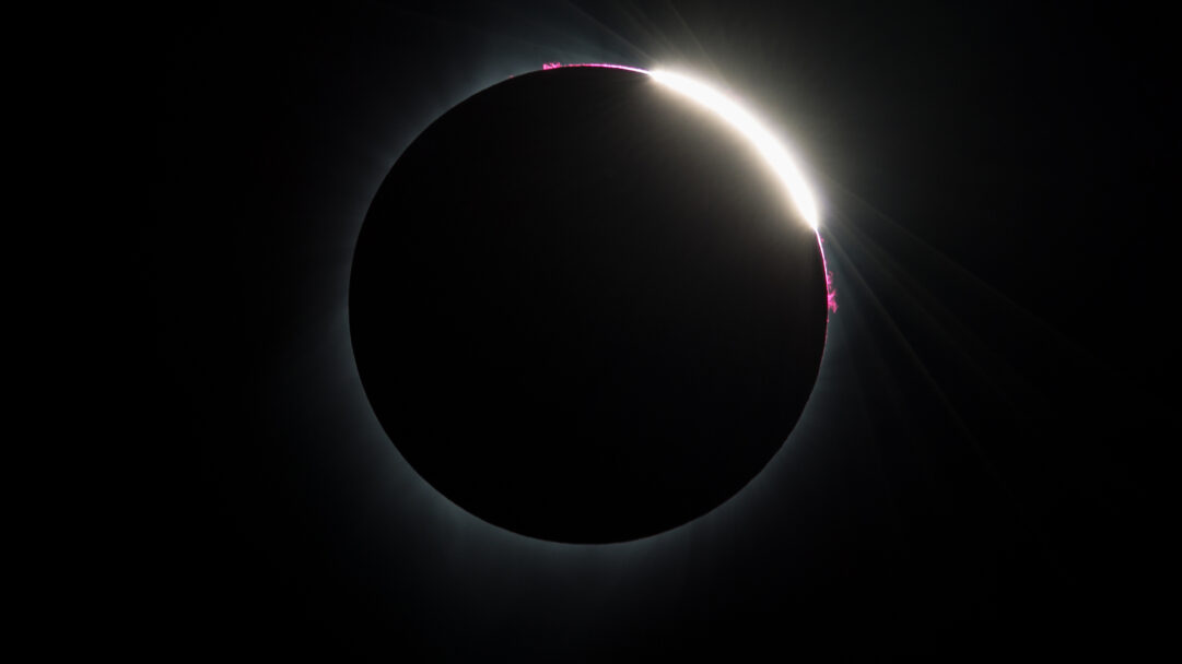 An awe-inspiring 4K wallpaper capturing the dramatic 'Diamond Ring' effect of the 2017 Total Solar Eclipse as seen from Madras, Oregon. The moon perfectly obscures the sun, revealing a single dazzling point of sunlight and a thin arc of vibrant pink solar prominences against the deep black sky.