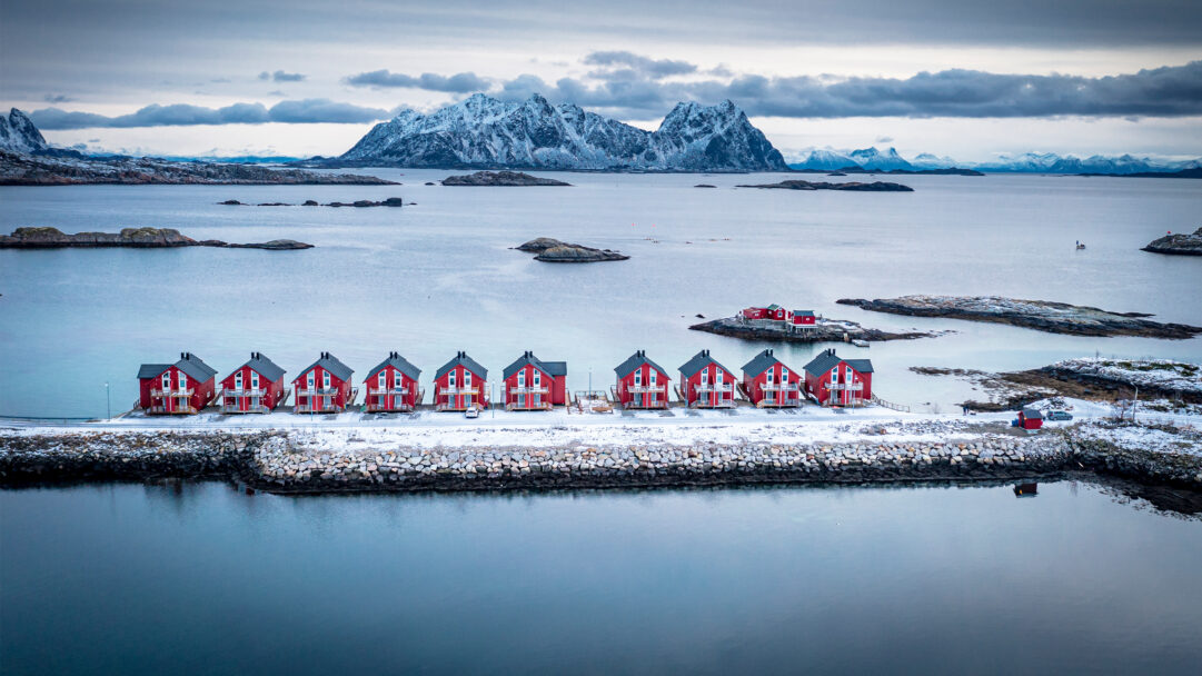 An iconic 4K wallpaper showcases the traditional red Rorbu houses of Svolvaer, Lofoten, Norway, set against a stunning winter landscape of a tranquil sea and snow-capped mountains. The vibrant crimson of the houses offers a striking contrast to the cool, muted blues and grays of the surrounding icy waters, rocky islets, and distant peaks, evoking a feeling of stark beauty and quiet resilience.