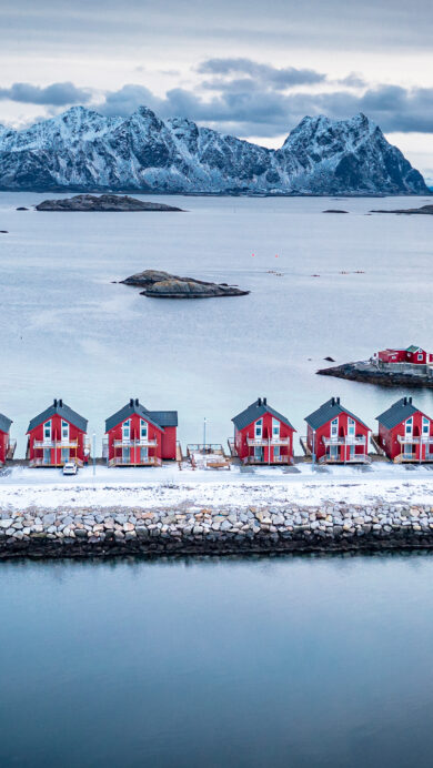 An iconic 4K wallpaper showcases the traditional red Rorbu houses of Svolvaer, Lofoten, Norway, set against a stunning winter landscape of a tranquil sea and snow-capped mountains. The vibrant crimson of the houses offers a striking contrast to the cool, muted blues and grays of the surrounding icy waters, rocky islets, and distant peaks, evoking a feeling of stark beauty and quiet resilience.
