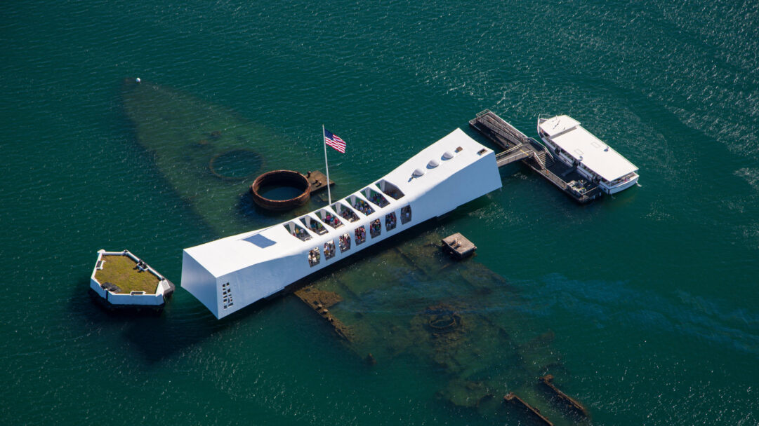 An evocative 4K wallpaper depicting the solemn USS Arizona Memorial in an aerial view over the tranquil waters of Pearl Harbor, Oahu, Hawaii. The pristine white memorial bridge sharply contrasts with the dark, discernible outline of the sunken battleship beneath the shimmering teal water, creating a deeply poignant scene of remembrance.