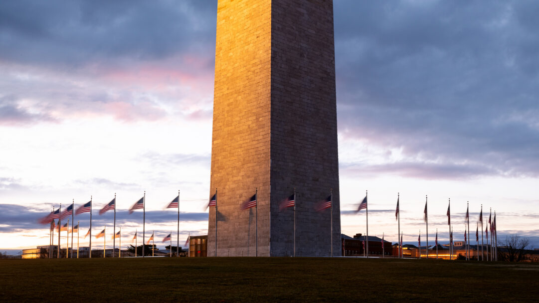 A vibrant 4K wallpaper showcasing the Washington Monument at sunset in Washington D.C., with a solemn array of American flags lining its base. The monument's stonework is bathed in the warm, golden light of the setting sun, creating a poignant contrast with the deepening twilight sky above and the subtle movement of the flags.