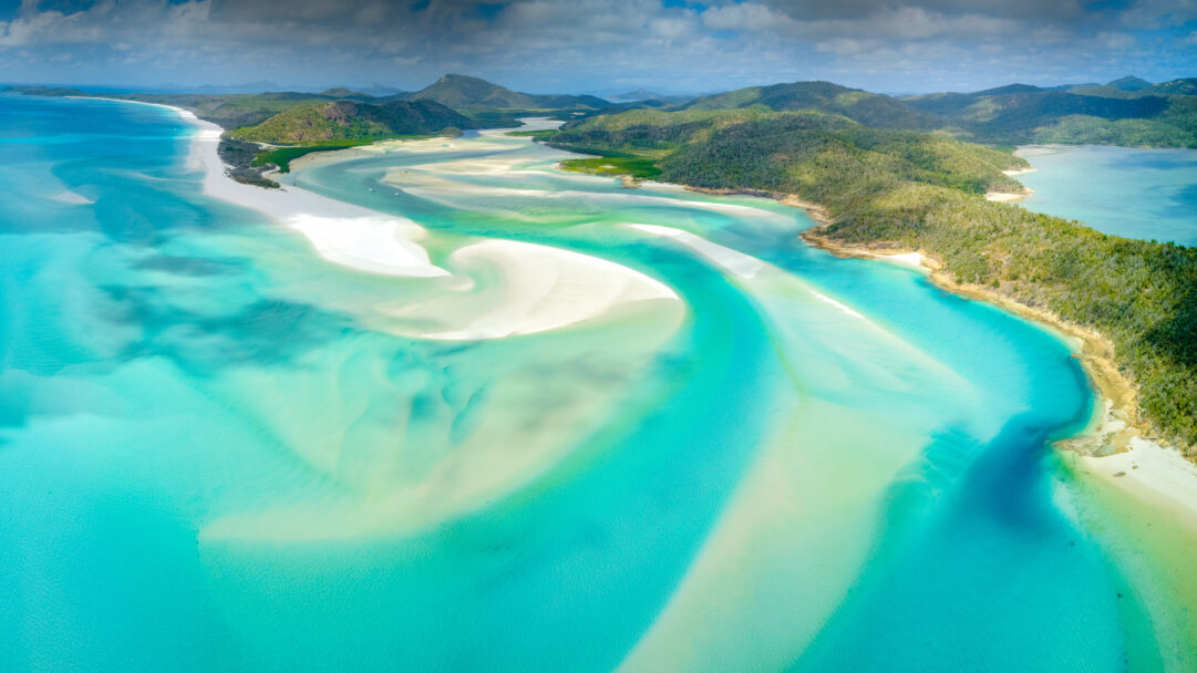 A magnificent 4K wallpaper captures an aerial view of the iconic Whitehaven Beach on Whitsunday Island, Queensland, Australia, curving between lush green hills and the Coral Sea. The pristine white silica sand creates stunning swirling patterns as it meets the multiple shades of vibrant turquoise and aquamarine waters, crafting a serene, dreamlike tropical landscape.