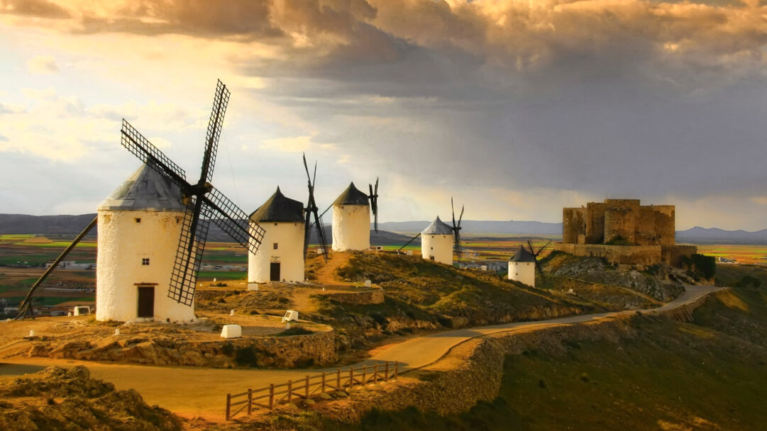 A dramatic 4K wallpaper unveils multiple traditional white windmills and a rugged stone castle atop a sun-drenched hill in Castilla-La Mancha, Spain. The landscape is bathed in the warm, golden light of sunset, sharply contrasting with dark, dramatic clouds overhead, creating a powerful and historic atmosphere.