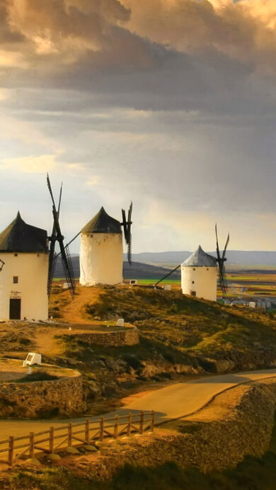 A dramatic 4K wallpaper unveils multiple traditional white windmills and a rugged stone castle atop a sun-drenched hill in Castilla-La Mancha, Spain. The landscape is bathed in the warm, golden light of sunset, sharply contrasting with dark, dramatic clouds overhead, creating a powerful and historic atmosphere.
