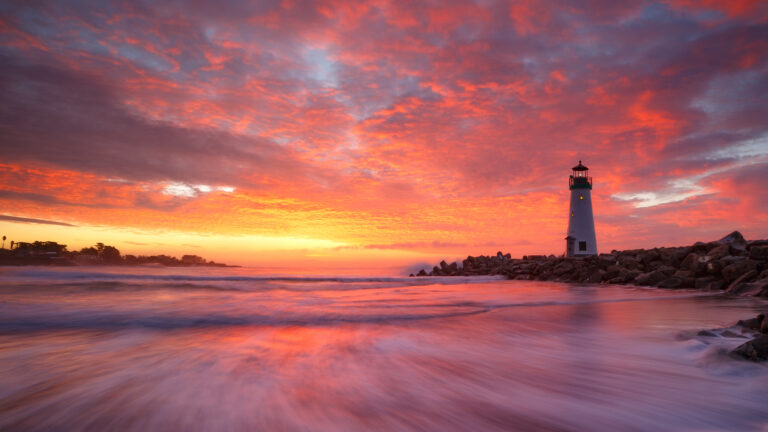 An awe-inspiring 4K wallpaper features the iconic Walton Lighthouse in Santa Cruz, California, standing against a fiery winter sunrise over the Pacific Ocean. The sky explodes with intense oranges and reds, casting a vibrant, painterly reflection across the long-exposure waves that sweep softly onto the rocky shore, creating a serene yet powerful scene.