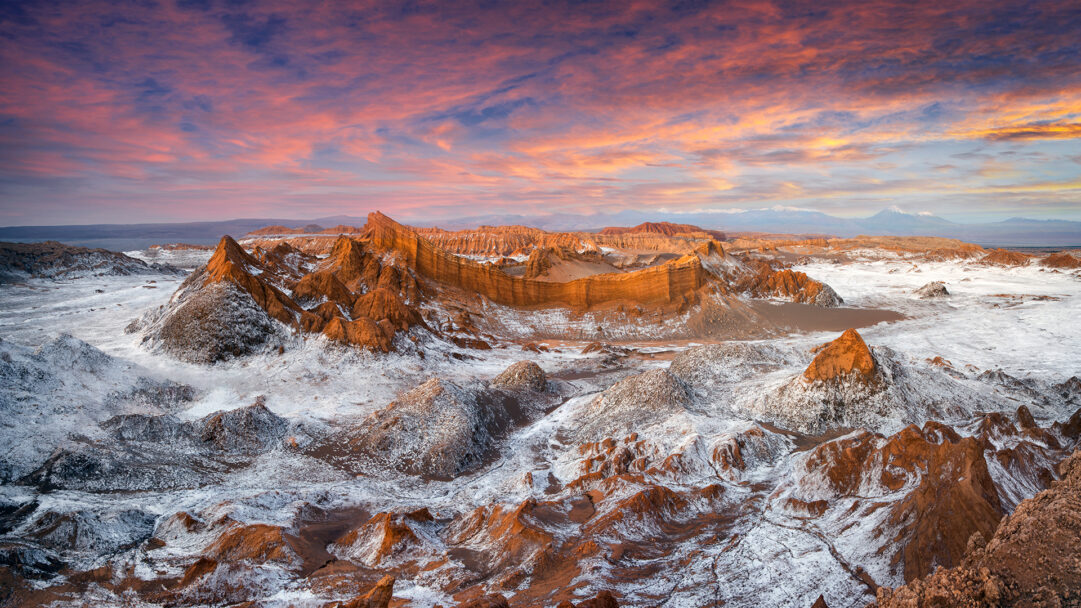 A breathtaking 4K wallpaper showcases the expansive desert landscape of Achaches Lookout in El Valle de la Luna, Chile, featuring dramatic, eroded rock formations at sunset. The sky's vibrant orange and pink clouds cast a warm glow on the reddish-brown peaks, sharply contrasting with the extensive white salt deposits that sculpt the valley floor, creating an otherworldly scene.