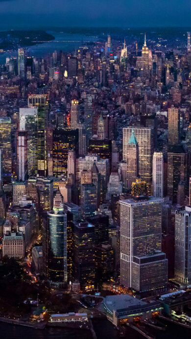An expansive 4K wallpaper showcasing an aerial view of the iconic Manhattan, New York City skyline at night. The glittering grid of countless building lights and illuminated bridges transforms the cityscape into a vibrant mosaic against the deep twilight sky and reflective dark waters.