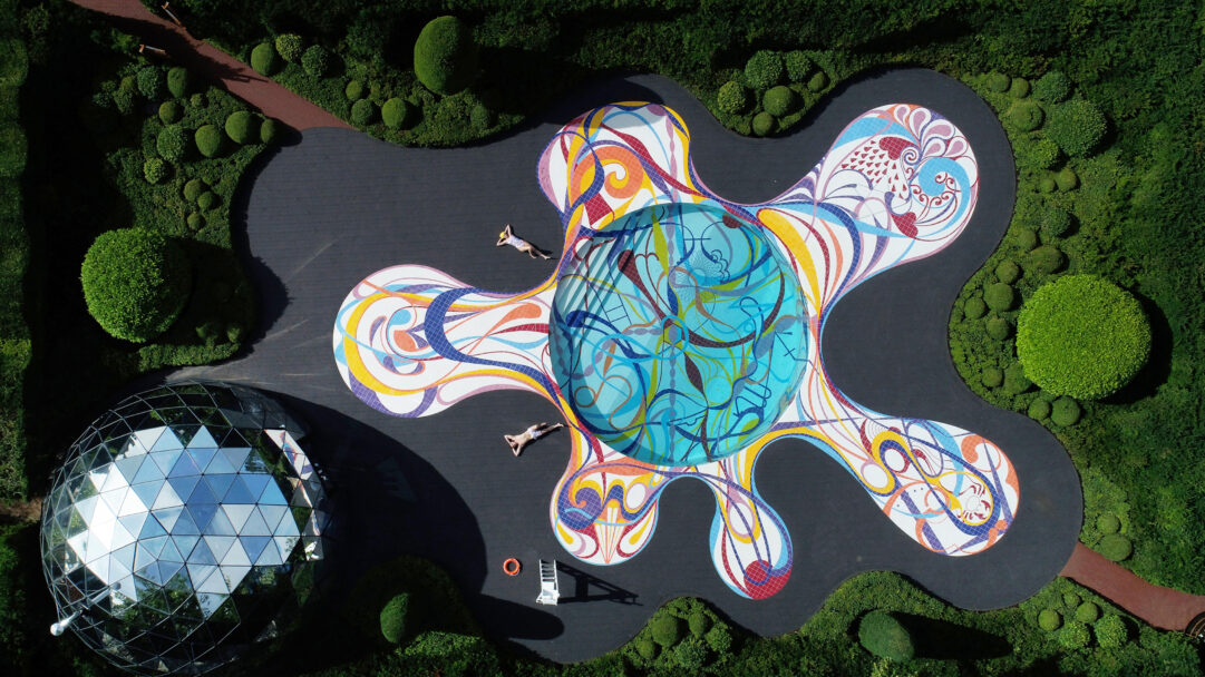 A captivating 4K wallpaper showcases an aerial park gateway landscape, featuring a vibrant, organically shaped abstract mosaic and a striking, reflective geodesic dome. The abstract mosaic's expansive, fluid, multi-colored patterns strikingly contrast with the dark paths and surrounding sculpted green topiaries, while the geodesic dome's mirrored facets reflect the sky, defining a mood of contemporary artistry and serene leisure within the park.