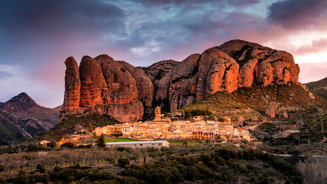 A majestic 4K wallpaper portraying Agüero, a Spanish village nestled beneath imposing rock formations in a vast, picturesque landscape. The scene is bathed in the warm, golden-orange light of a sunset, dramatically highlighting the vertical cliffs and infusing the sky with hues of deep blue and soft pink.