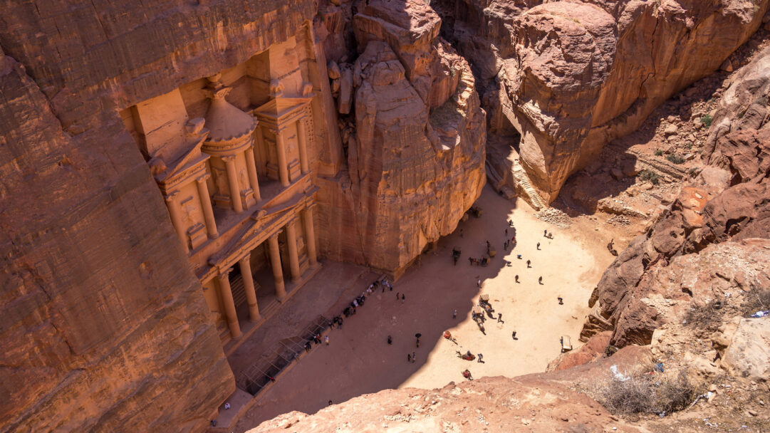 An iconic 4K wallpaper showcases Al-Khazneh, The Treasury of Petra, Jordan, deeply carved into the towering, reddish sandstone cliffs of a desert canyon. The dramatic interplay of strong sunlight and deep shadows accentuates the intricate façade's details and the vast scale of the ancient monument against the bustling ground of small figures.