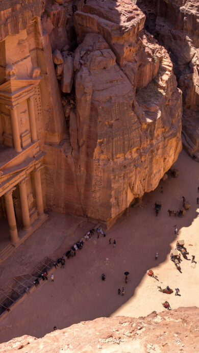 An iconic 4K wallpaper showcases Al-Khazneh, The Treasury of Petra, Jordan, deeply carved into the towering, reddish sandstone cliffs of a desert canyon. The dramatic interplay of strong sunlight and deep shadows accentuates the intricate façade's details and the vast scale of the ancient monument against the bustling ground of small figures.