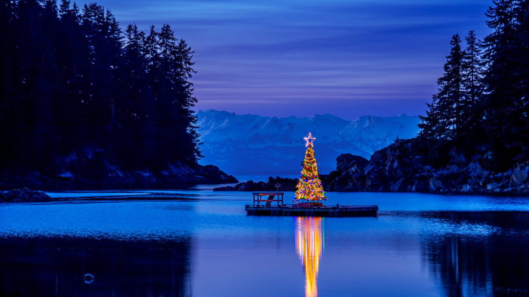 A magical 4K wallpaper features a brightly lit Christmas tree floating on a tranquil body of water in Amalga Harbor, Alaska, flanked by dark, forested shores and distant snowy mountains under the twilight sky. Its vibrant, multicolored lights cast a stunning, elongated reflection that shimmers across the deep blue surface, creating a captivating interplay of festive warmth and serene coolness.