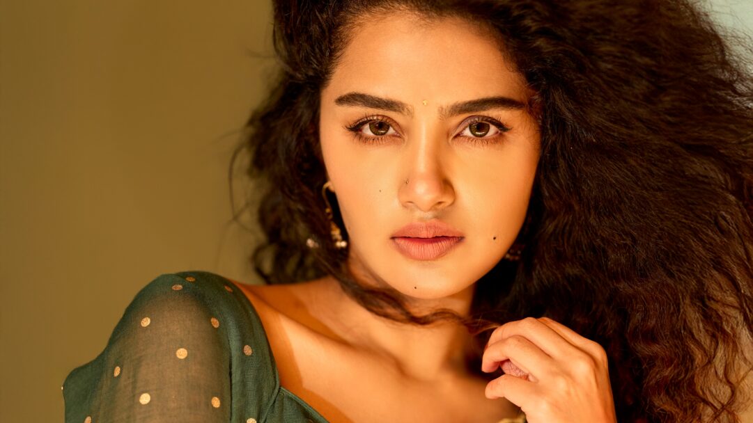 A captivating 4K wallpaper of Anupama Parameswaran in a close-up portrait, looking directly at the viewer with a gentle gaze against a warm, soft background. Her dark, voluminous curly hair contrasts beautifully with her glowing skin, enhanced by warm, soft lighting that highlights her serene expression and the delicate details of her eyes and traditional nose stud.