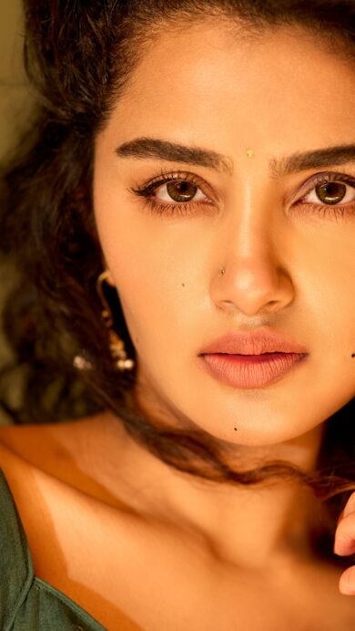 A captivating 4K wallpaper of Anupama Parameswaran in a close-up portrait, looking directly at the viewer with a gentle gaze against a warm, soft background. Her dark, voluminous curly hair contrasts beautifully with her glowing skin, enhanced by warm, soft lighting that highlights her serene expression and the delicate details of her eyes and traditional nose stud.