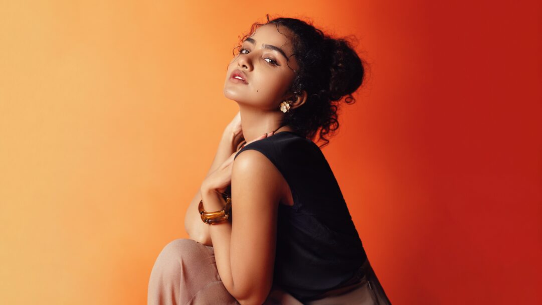 A radiant 4K wallpaper displays Anupama Parameswaran in a captivating photoshoot, posing confidently against a striking orange-red gradient background. Her elegant pose and direct gaze are beautifully illuminated by the warm, transitioning colors, creating a vibrant and sophisticated mood.