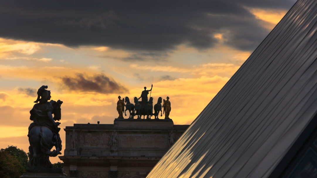 A majestic 4K wallpaper of the Arc de Triomphe du Carrousel and the Louvre Pyramid in Paris, dramatically silhouetted against a sunset sky. The captivating interplay of light showcases their dark forms against the vibrant golden-orange sunset, dramatically contrasted by a band of dark, heavy clouds.