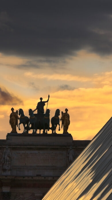 A majestic 4K wallpaper of the Arc de Triomphe du Carrousel and the Louvre Pyramid in Paris, dramatically silhouetted against a sunset sky. The captivating interplay of light showcases their dark forms against the vibrant golden-orange sunset, dramatically contrasted by a band of dark, heavy clouds.