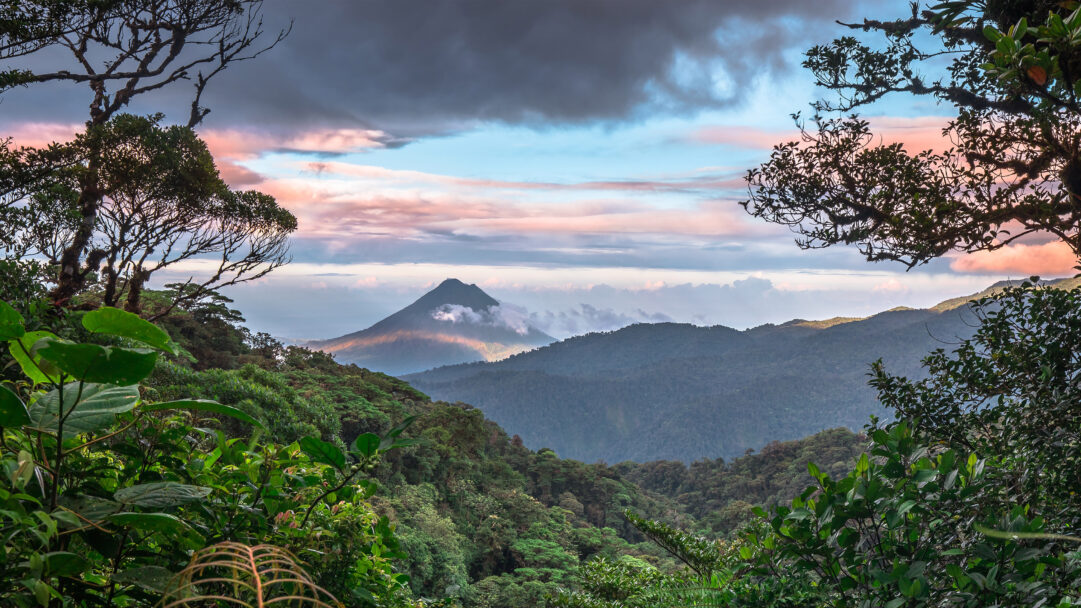 An awe-inspiring 4K wallpaper showcasing the iconic Arenal Volcano towering over a lush, misty landscape as viewed from Monteverde, Costa Rica. Streaks of sunlight illuminate the volcano's misty lower slopes, contrasting with the rich green rainforest canopy, all under a captivating sky of soft pink and grey-blue clouds.