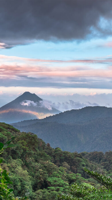 An awe-inspiring 4K wallpaper showcasing the iconic Arenal Volcano towering over a lush, misty landscape as viewed from Monteverde, Costa Rica. Streaks of sunlight illuminate the volcano's misty lower slopes, contrasting with the rich green rainforest canopy, all under a captivating sky of soft pink and grey-blue clouds.