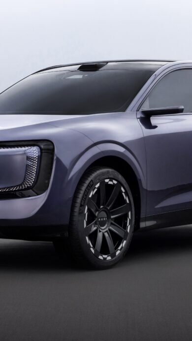 A visionary 4K wallpaper presenting a futuristic Audi E-SUV concept car, posed elegantly on a dark grey surface against a bright, minimalist backdrop. Its subtle purple-grey metallic sheen and the distinctive geometric light patterns on its front grille create a sophisticated and modern aesthetic.