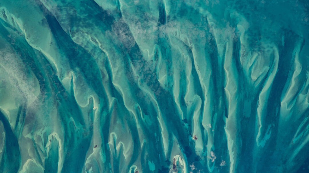 An expansive 4K wallpaper revealing the mesmerizing blue-green ocean waters of the Bahamas, as viewed from the International Space Station, where shallow sandbanks and deeper channels create a tapestry of natural patterns. Varying depths are accentuated by an exquisite interplay of turquoise, aqua, and deep teal hues, highlighting the intricate underwater topography.