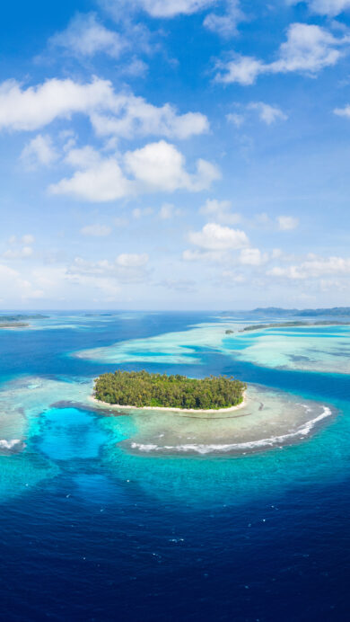 A captivating 4K wallpaper showcases the Banyak Islands of Sumatra, Indonesia, from an aerial view across a sprawling tropical ocean. The scene is defined by the mesmerizing gradient of deep blue to clear turquoise waters revealing intricate coral reefs, encircling lush, palm-covered islets beneath a bright, cloud-scattered sky.