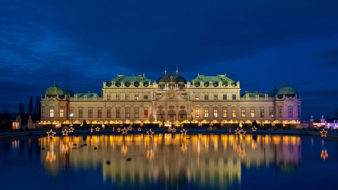 A magical 4K wallpaper showcasing the illuminated Belvedere Palace at night, set against the enchanting backdrop of its Vienna Christmas Market. Its festive lights, along with glowing star decorations, create a breathtaking, vibrant reflection on the calm water, capturing a serene holiday atmosphere.