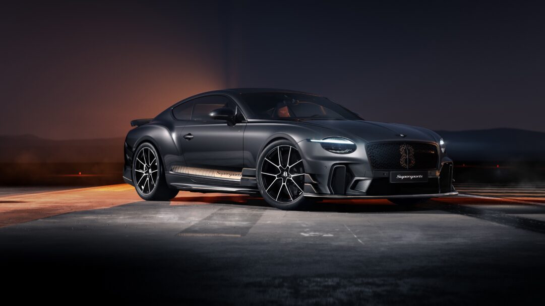 A sophisticated 4K wallpaper showcasing a dark gray Bentley Continental GT Supersports coupe parked on an asphalt surface at dusk. Its bright LED headlights cut through the dramatic low light, enhanced by warm orange reflections across the ground and a distant, dusky horizon.