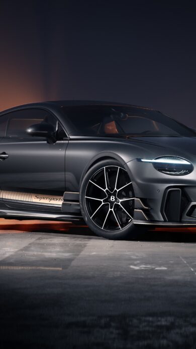 A sophisticated 4K wallpaper showcasing a dark gray Bentley Continental GT Supersports coupe parked on an asphalt surface at dusk. Its bright LED headlights cut through the dramatic low light, enhanced by warm orange reflections across the ground and a distant, dusky horizon.