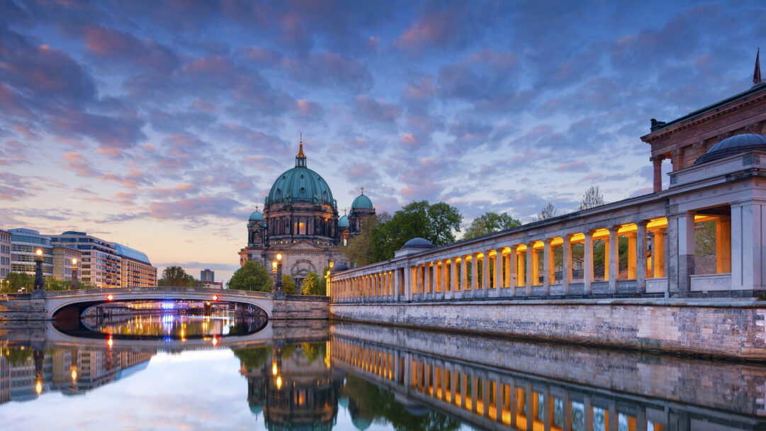 A breathtaking 4K wallpaper illustrates the magnificent Berlin Cathedral and Museum Island situated along a tranquil river in Berlin, Germany, as dusk settles. Warm lights from the surrounding buildings and a bridge glow brilliantly, mirroring their colors in the smooth water below under a sky painted with soft twilight hues.