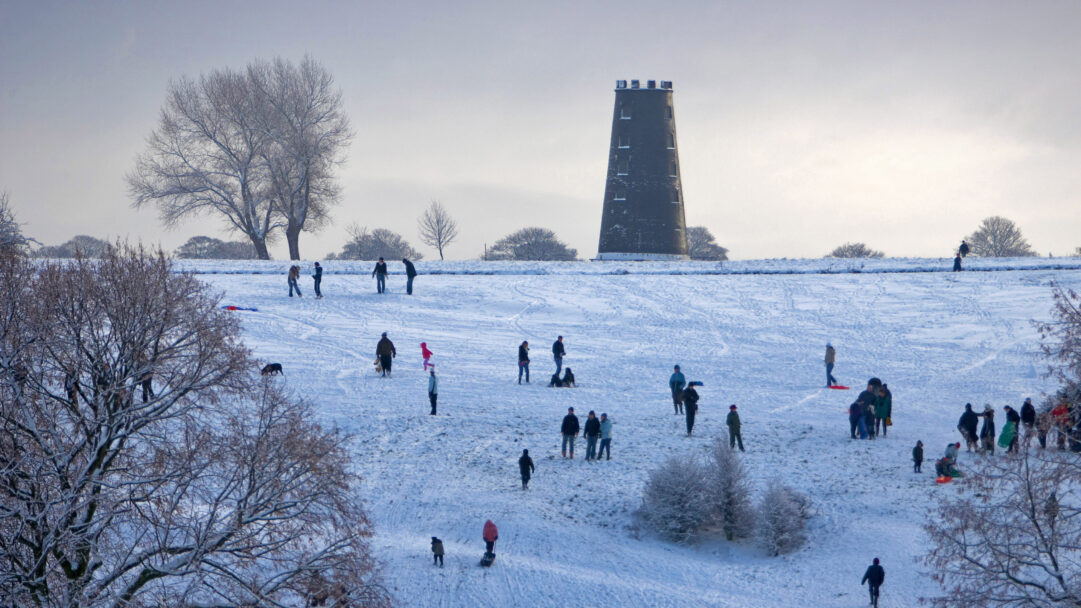 A captivating 4K wallpaper captures the joyous winter scene of people sledding down snow-covered slopes on Beverley Westwood Common, with a historic tower silhouetted against the horizon. The pristine white snow, crisscrossed with sled tracks and footprints, creates a vivid contrast with the dark, sturdy form of the tower and the skeletal branches of nearby trees, evoking a cheerful winter's day.