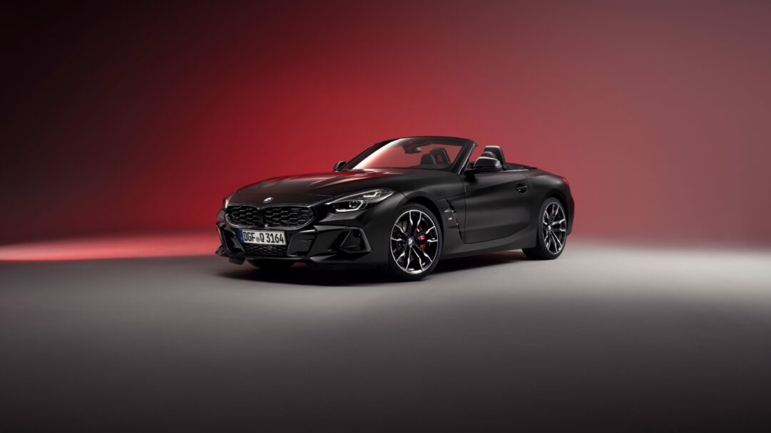 A dynamic 4K wallpaper featuring a sleek black BMW Z4 Roadster sports car positioned on a dark grey surface against a vibrant red background. The car's aggressive lines and subtle red brake calipers pop against the radiant, gradient red backdrop, creating an intense and luxurious mood.