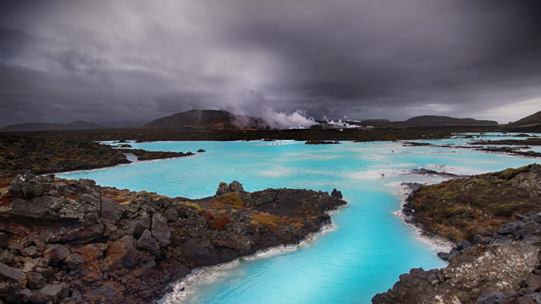A striking 4K wallpaper reveals the iconic Blue Lagoon in Grindavík, Iceland, as a mesmerizing geothermal landscape of vivid turquoise pools. Dark volcanic rock formations line the vibrant water, while plumes of steam rise dramatically from a distant geothermal plant under a brooding, cloudy sky.