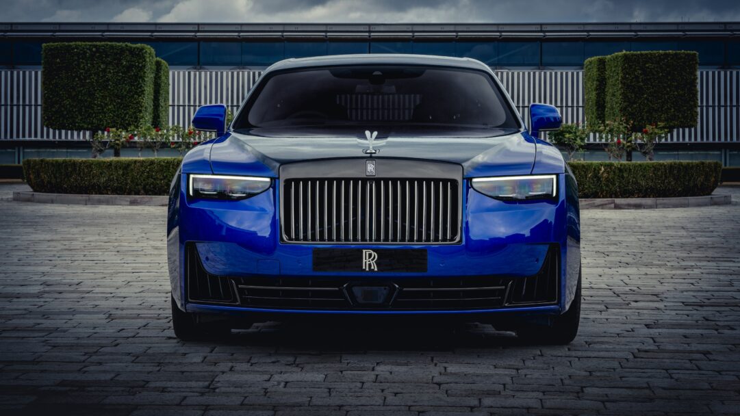 An exquisite 4K wallpaper showcasing a majestic blue Rolls-Royce Ghost luxury car, positioned in a commanding front view on a paved surface before a modern building and neatly trimmed hedges under an overcast sky. The car's brilliant headlights and polished deep blue finish powerfully reflect the muted light, underscoring its imposing elegance.