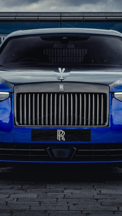 An exquisite 4K wallpaper showcasing a majestic blue Rolls-Royce Ghost luxury car, positioned in a commanding front view on a paved surface before a modern building and neatly trimmed hedges under an overcast sky. The car's brilliant headlights and polished deep blue finish powerfully reflect the muted light, underscoring its imposing elegance.