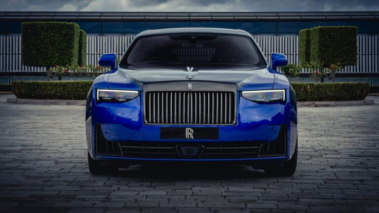 An exquisite 4K wallpaper showcasing a majestic blue Rolls-Royce Ghost luxury car, positioned in a commanding front view on a paved surface before a modern building and neatly trimmed hedges under an overcast sky. The car's brilliant headlights and polished deep blue finish powerfully reflect the muted light, underscoring its imposing elegance.