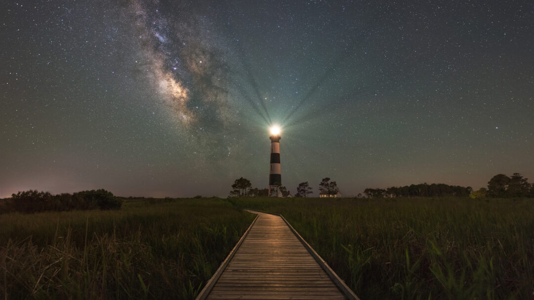 An enchanting 4K wallpaper presenting the majestic Bodie Island Lighthouse in Nags Head, North Carolina, standing tall under a vast night sky. Its powerful beam pierces the dark, starry expanse, beautifully highlighting the glowing band of the Milky Way stretching across the heavens.