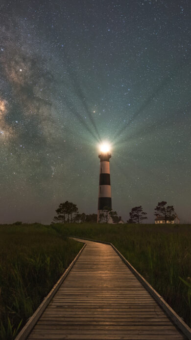 An enchanting 4K wallpaper presenting the majestic Bodie Island Lighthouse in Nags Head, North Carolina, standing tall under a vast night sky. Its powerful beam pierces the dark, starry expanse, beautifully highlighting the glowing band of the Milky Way stretching across the heavens.
