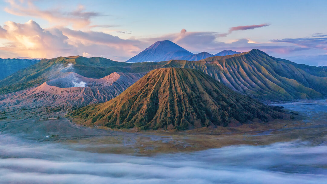 A breathtaking 4K wallpaper showcasing the majestic volcanic landscape of Bromo Tengger Semeru National Park in East Java, Indonesia. Wisps of mist gently blanket the foreground, while a smoking crater and towering peaks are bathed in the soft, warm glow of dawn under a sky streaked with pastel hues.