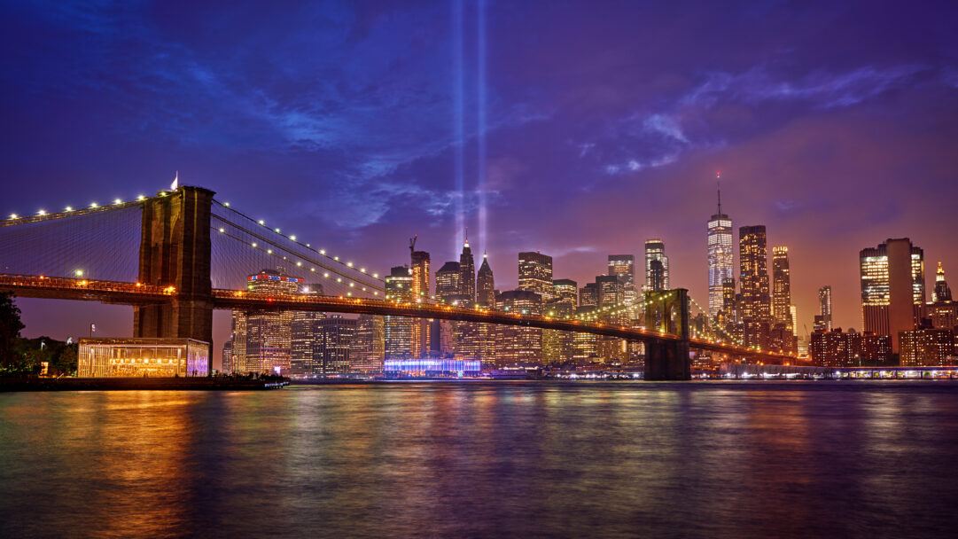 A poignant 4K wallpaper showcasing the iconic Brooklyn Bridge and the illuminated New York City skyline at night. Two powerful, luminous beams of blue light pierce the deep twilight sky, forming the 9/11 Tribute in Light, reflected alongside the golden city lights on the calm water below.