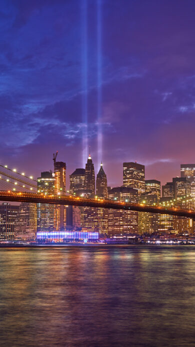 A poignant 4K wallpaper showcasing the iconic Brooklyn Bridge and the illuminated New York City skyline at night. Two powerful, luminous beams of blue light pierce the deep twilight sky, forming the 9/11 Tribute in Light, reflected alongside the golden city lights on the calm water below.