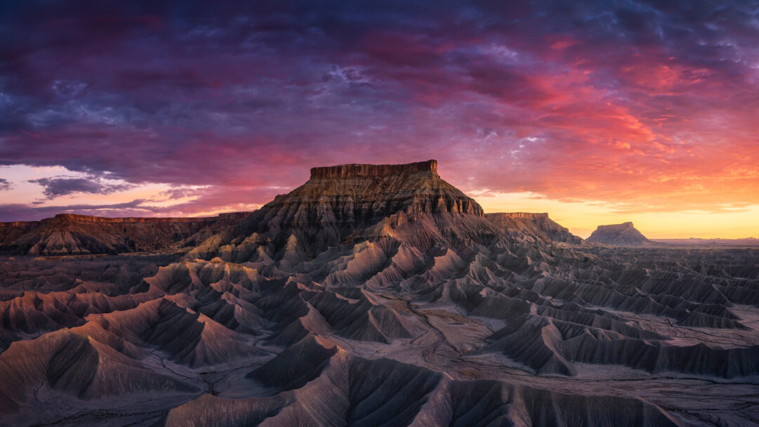 A breathtaking 4K wallpaper featuring a majestic sandstone butte towering over a vast, undulating badlands landscape in Capitol Reef National Park, Utah. The sky blazes with a vibrant sunset gradient of deep purples, fiery reds, and golden oranges, casting dramatic shadows and warm light across the rugged geological formations.