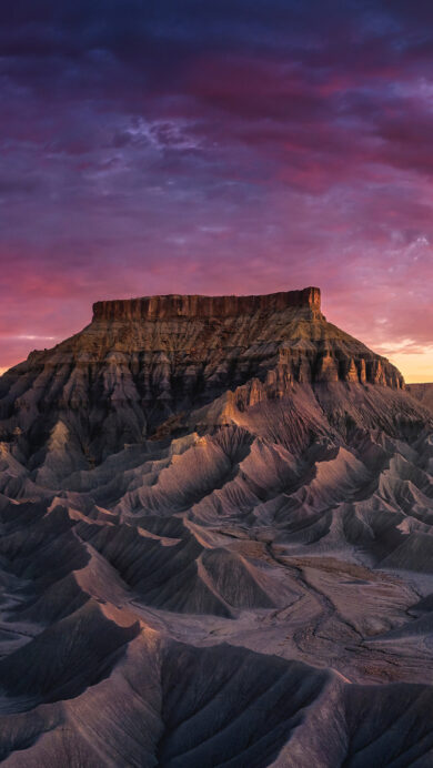 A breathtaking 4K wallpaper featuring a majestic sandstone butte towering over a vast, undulating badlands landscape in Capitol Reef National Park, Utah. The sky blazes with a vibrant sunset gradient of deep purples, fiery reds, and golden oranges, casting dramatic shadows and warm light across the rugged geological formations.