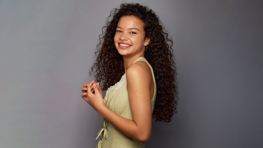 A captivating 4K wallpaper of Catherine Laga'aia in a vibrant studio portrait against a smooth gray background. Her radiant, open smile and rich cascade of dark, curly hair create an engaging and lively mood.