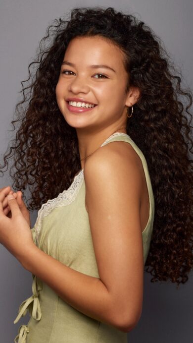 A captivating 4K wallpaper of Catherine Laga'aia in a vibrant studio portrait against a smooth gray background. Her radiant, open smile and rich cascade of dark, curly hair create an engaging and lively mood.