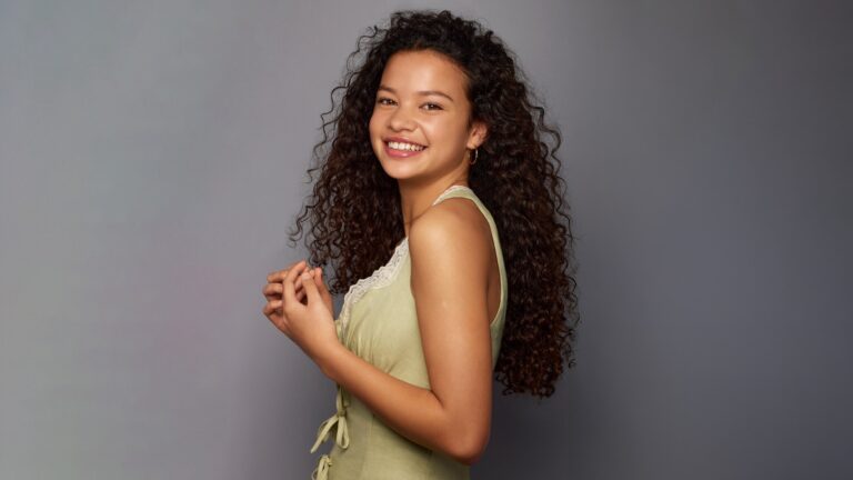 A captivating 4K wallpaper of Catherine Laga'aia in a vibrant studio portrait against a smooth gray background. Her radiant, open smile and rich cascade of dark, curly hair create an engaging and lively mood.