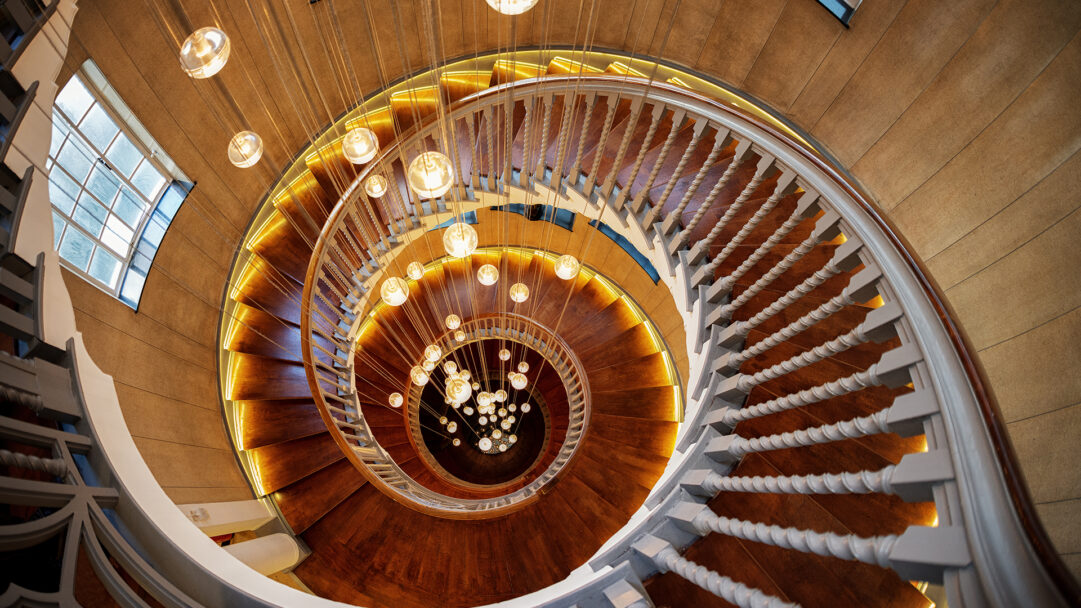 A captivating 4K wallpaper of the iconic Cecil Brewer Spiral Staircase in London's Heal's Building, viewed from above, reveals its stunning architectural form. The polished wooden steps are dramatically illuminated by a continuous warm glow, while a mesmerizing cascade of spherical pendant lights descends through the spiral's core, casting a magical golden ambiance.