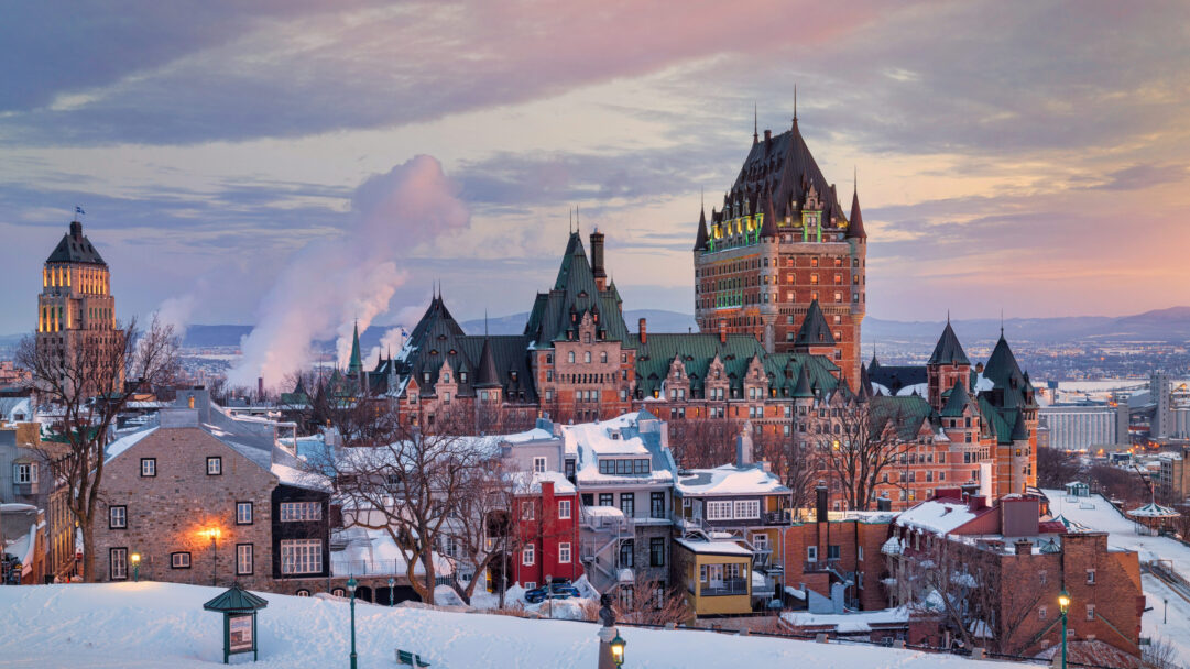 A captivating 4K wallpaper showcases the iconic Château Frontenac towering over a snow-covered Quebec City during a serene winter sunset. Its grand architecture is illuminated by a warm interior glow, beautifully contrasting with the soft pinks and purples of the fading sunset sky and the pristine white of the deep winter snow.