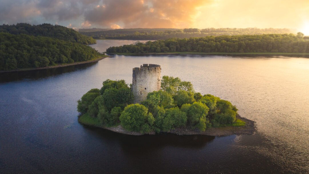 An evocative 4K wallpaper presenting Cloughoughter Castle, a historic stone ruin, situated on a small, lush island in Lough Oughter, Ireland, surrounded by dense forests. The warm, golden light of the sunset paints the clouds and the distant landscape, creating a majestic and peaceful ambiance that perfectly complements the ancient castle.