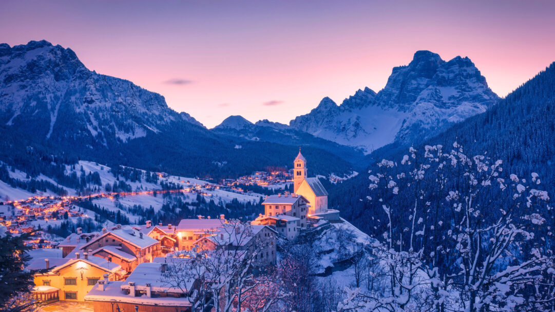 An ethereal 4K wallpaper showcasing the picturesque Colle Santa Lucia Village nestled among the snow-covered Dolomites in Italy. Warm lights from the snow-draped village homes glow invitingly, creating a magical contrast with the cool blues of the majestic, snow-capped Dolomites and the soft pink-purple sunset sky.